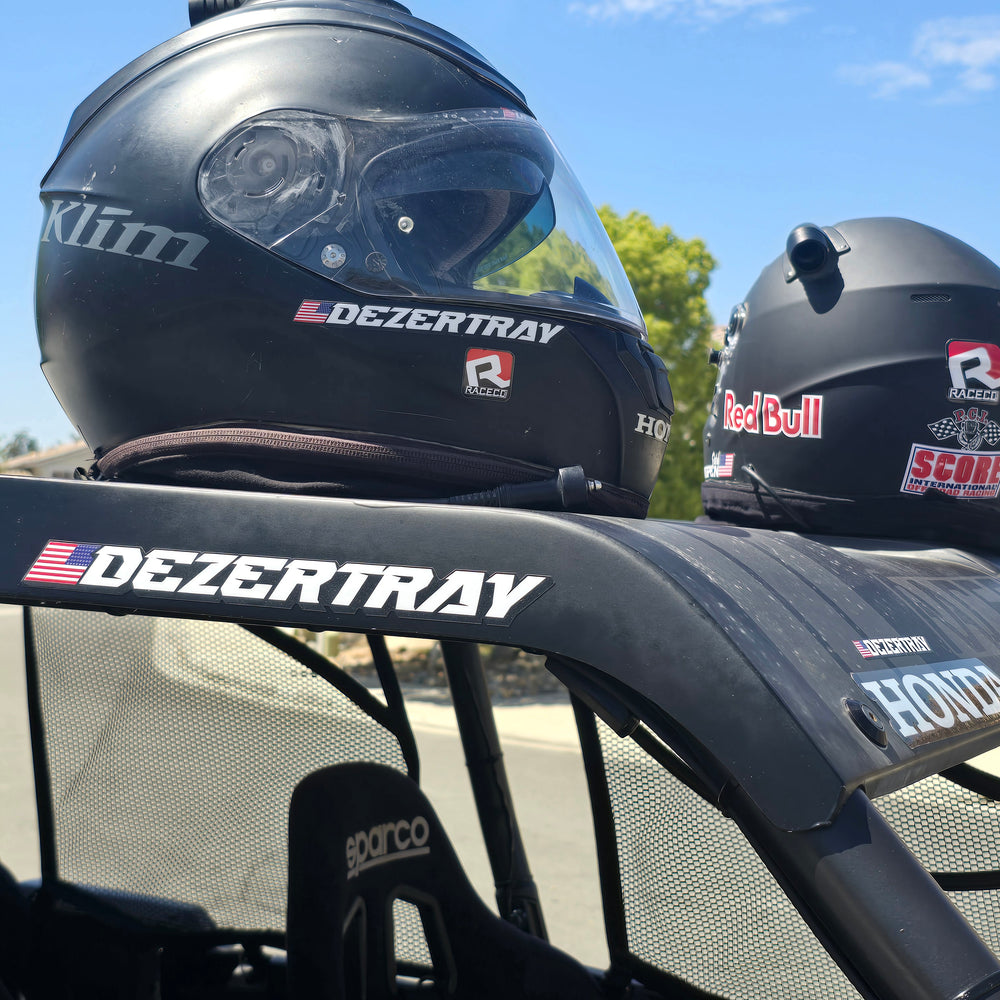 Close-up of personalized name and flag sticker on the roof panel of a UTV. Driver wearing a helmet with desert landscape in the background.