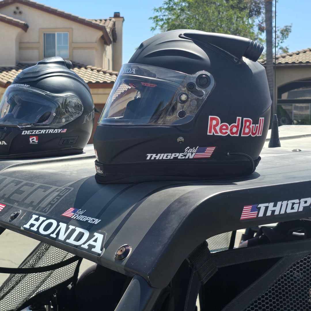 UTV parked under bright desert sun showing custom name and flag sticker on rear quarter panel. Driver and passenger wearing helmets.